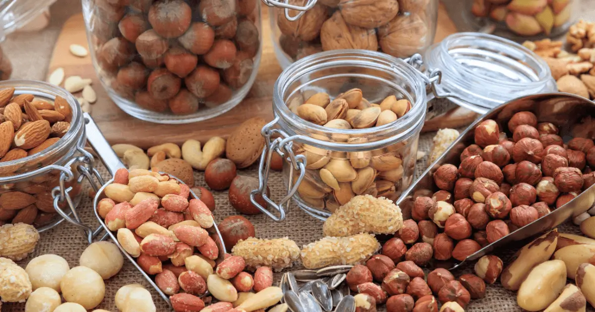 Mélange de fruits à coque en vrac Divers fruits à coque, comme les amandes, noisettes, pistaches, et cacahuètes, disposés dans des bocaux en verre et des pelles en métal sur une table en toile de jute.
